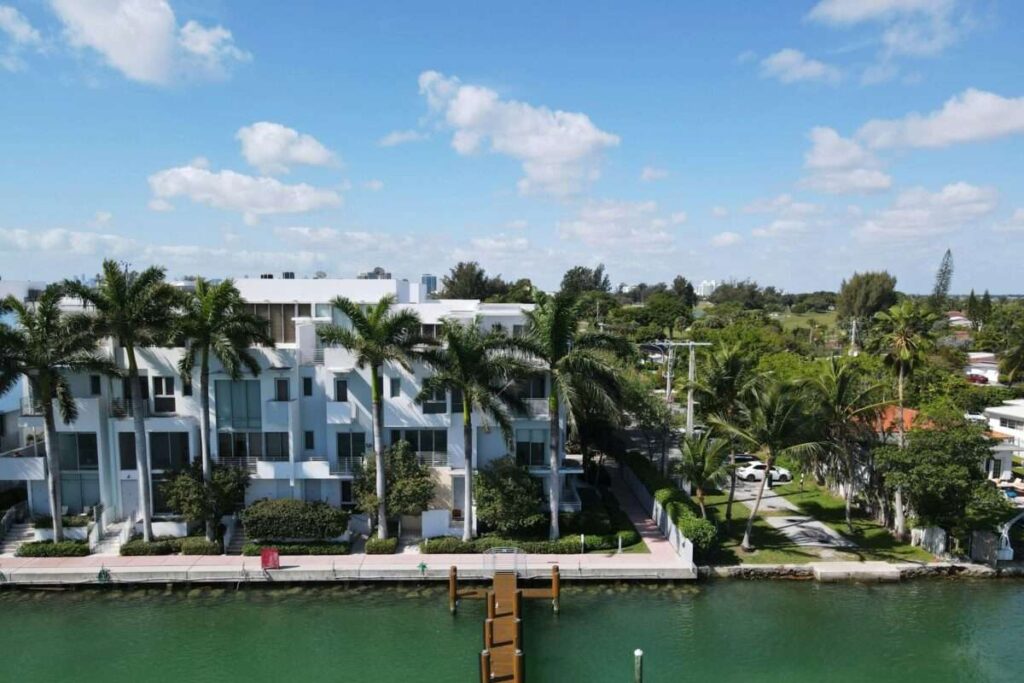 White modern apartment buildings line a canal, with tall palm trees and a bright blue sky above.