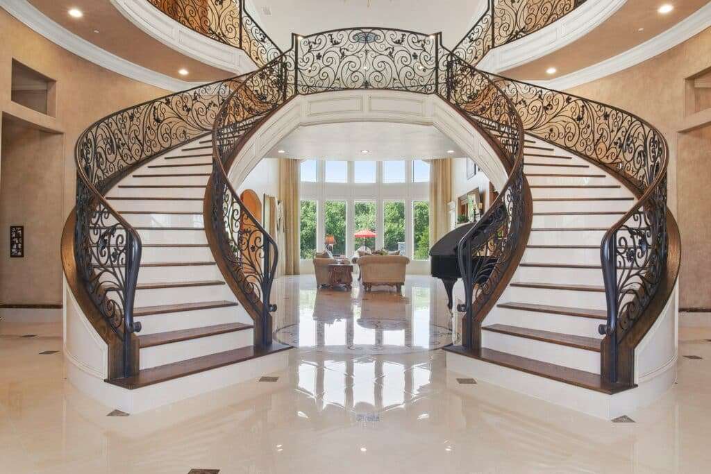 Grand two-sided staircase with ornate wrought-iron railing in a luxurious foyer; light beige walls and polished marble floor.