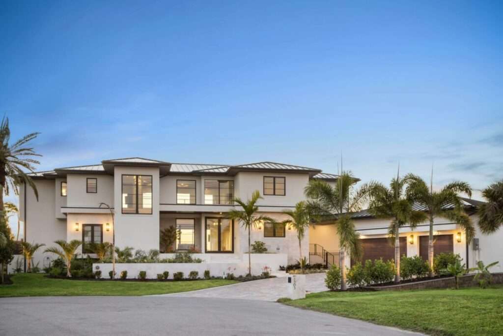 Modern two-story house with large windows, white exterior, and tropical landscaping under a blue sky.