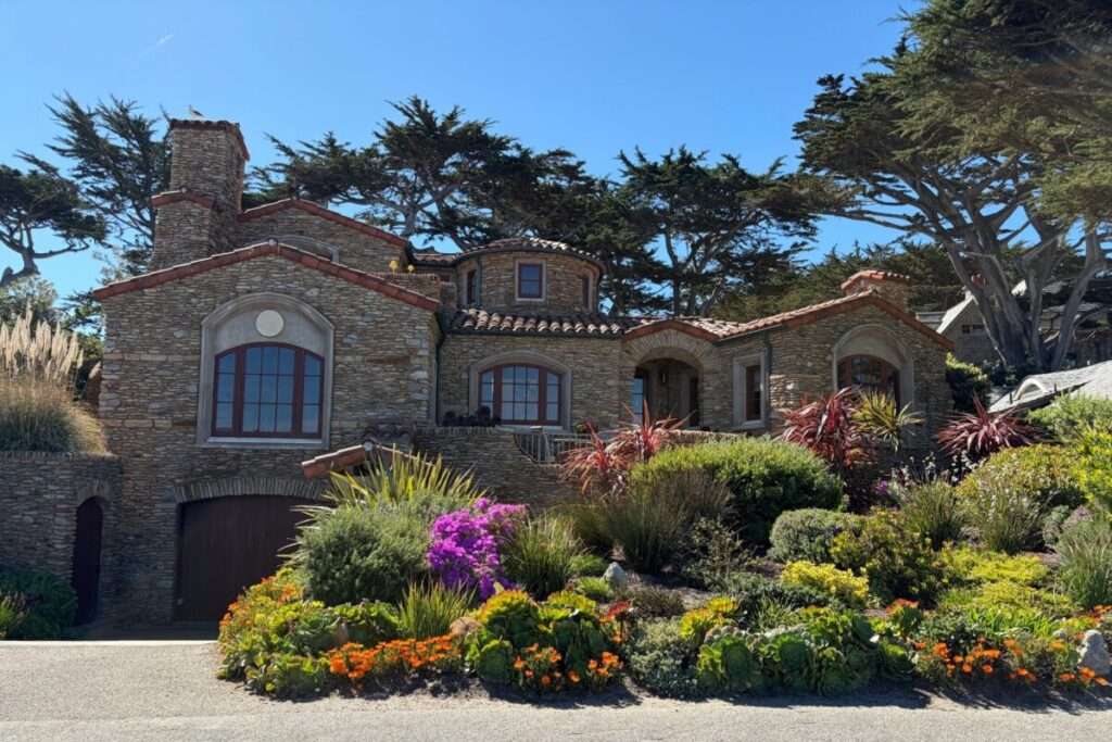 Stone house with rounded tower, red tile roof, and arched windows, set behind vibrant desert garden and blue sky.