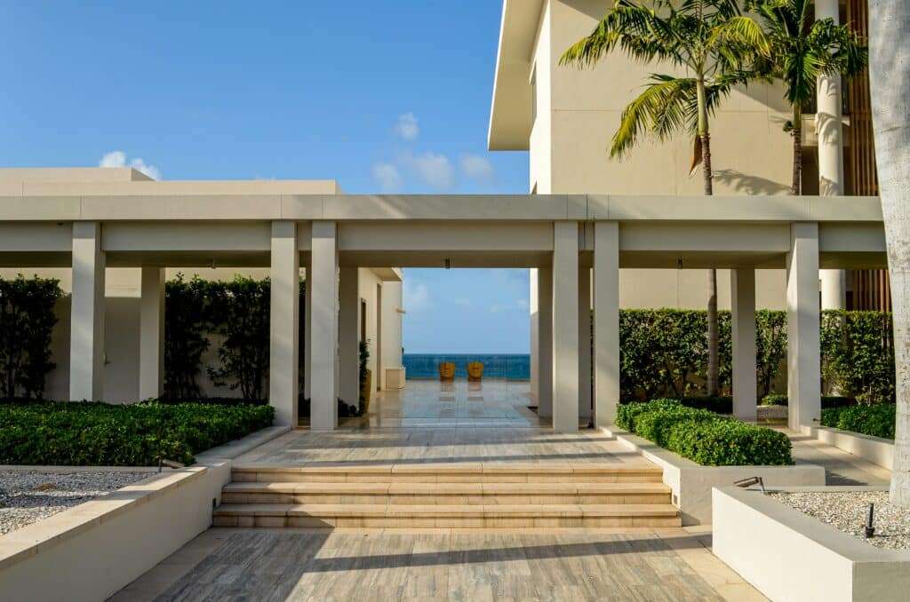 Beige modern courtyard with white pillars forming a covered walkway, leading to a view of the blue sea between the columns, flanked by palm trees and steps in the foreground.