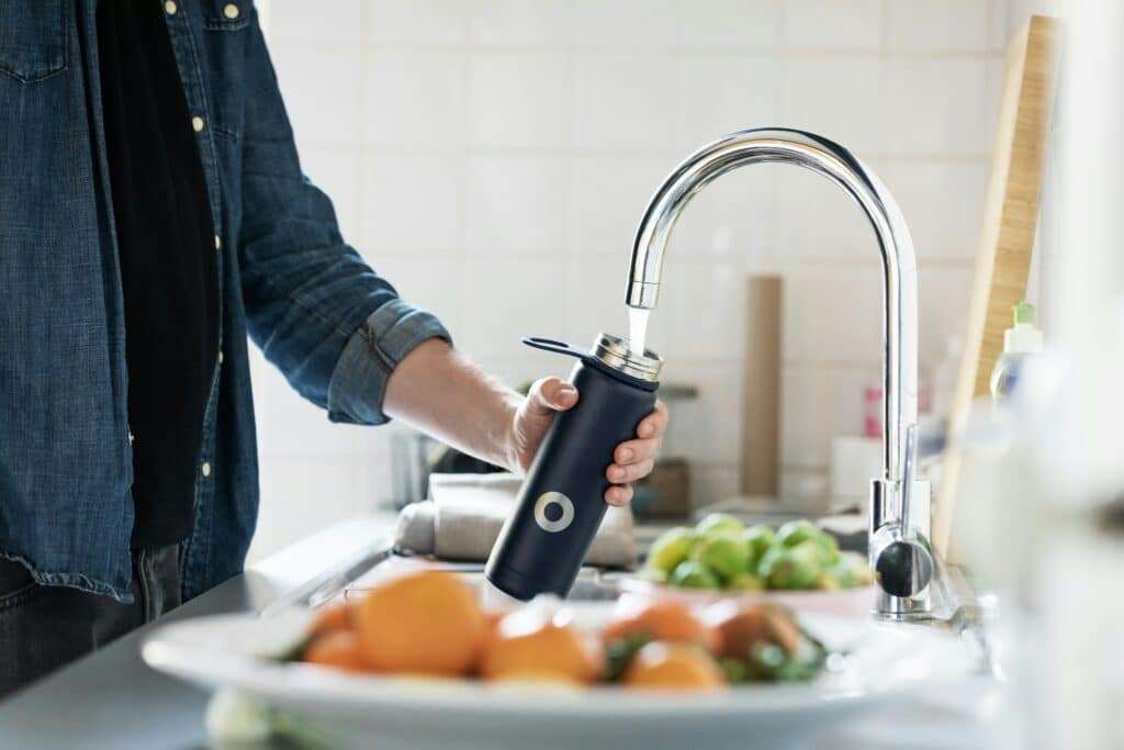 Person in a denim shirt fills a dark reusable bottle at a modern kitchen sink under running water.