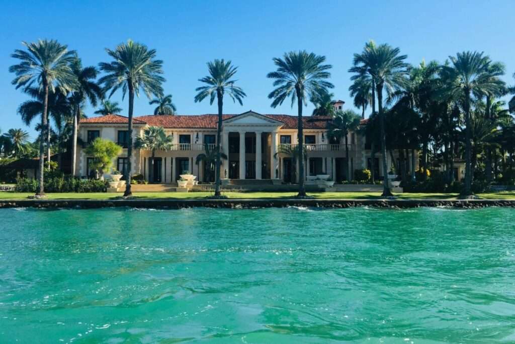 Waterfront villa with red-tiled roof framed by tall palm trees on a sunny day, turquoise water in the foreground