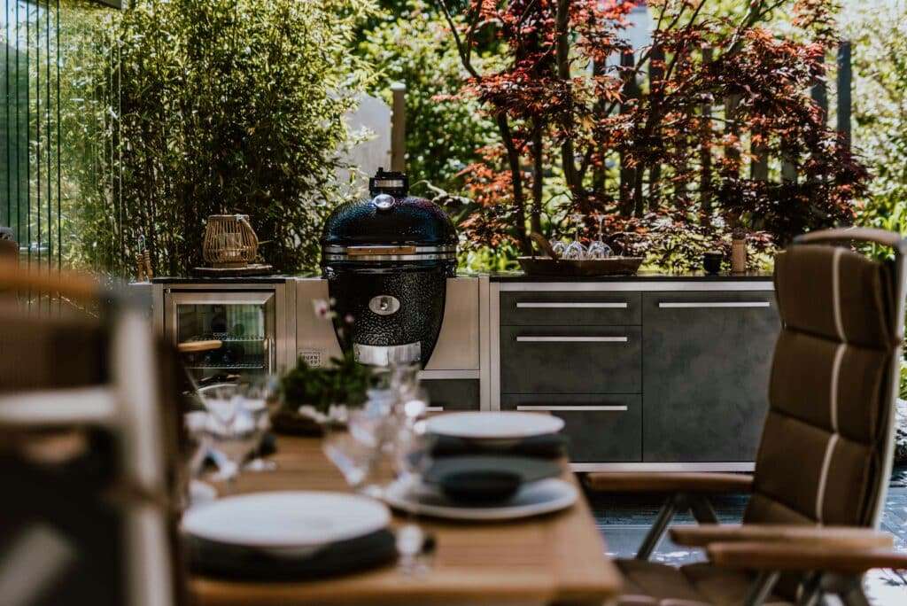 Outdoor kitchen with a black Big Green Egg kamado grill on a stainless counter, surrounded by greenery.