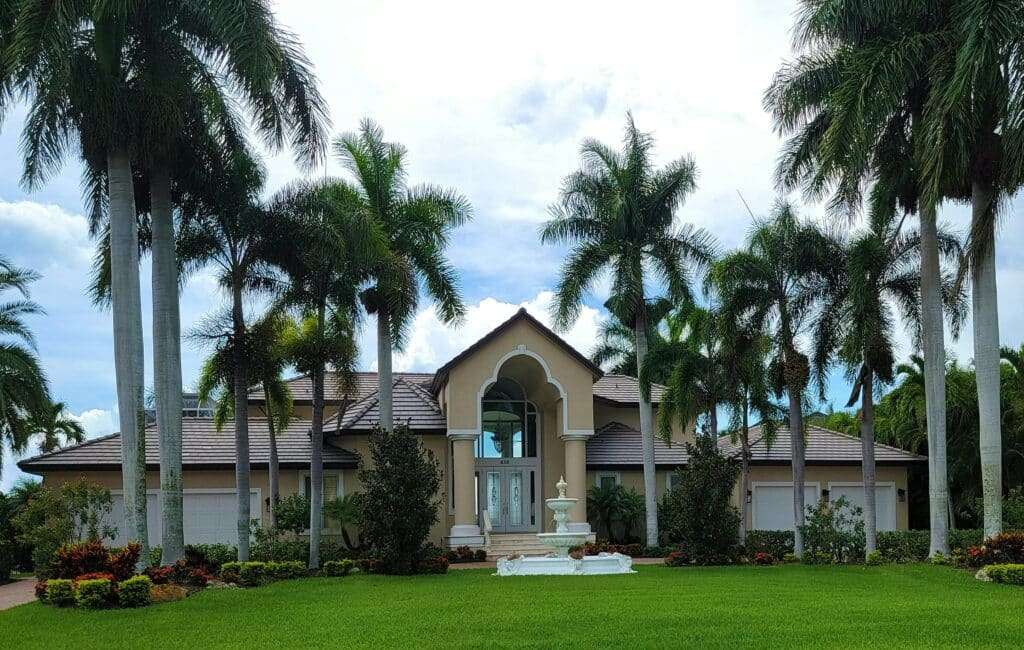 A beige tropical-style house with a centered archway, surrounded by tall palm trees and a manicured green lawn with a white fountain in front.