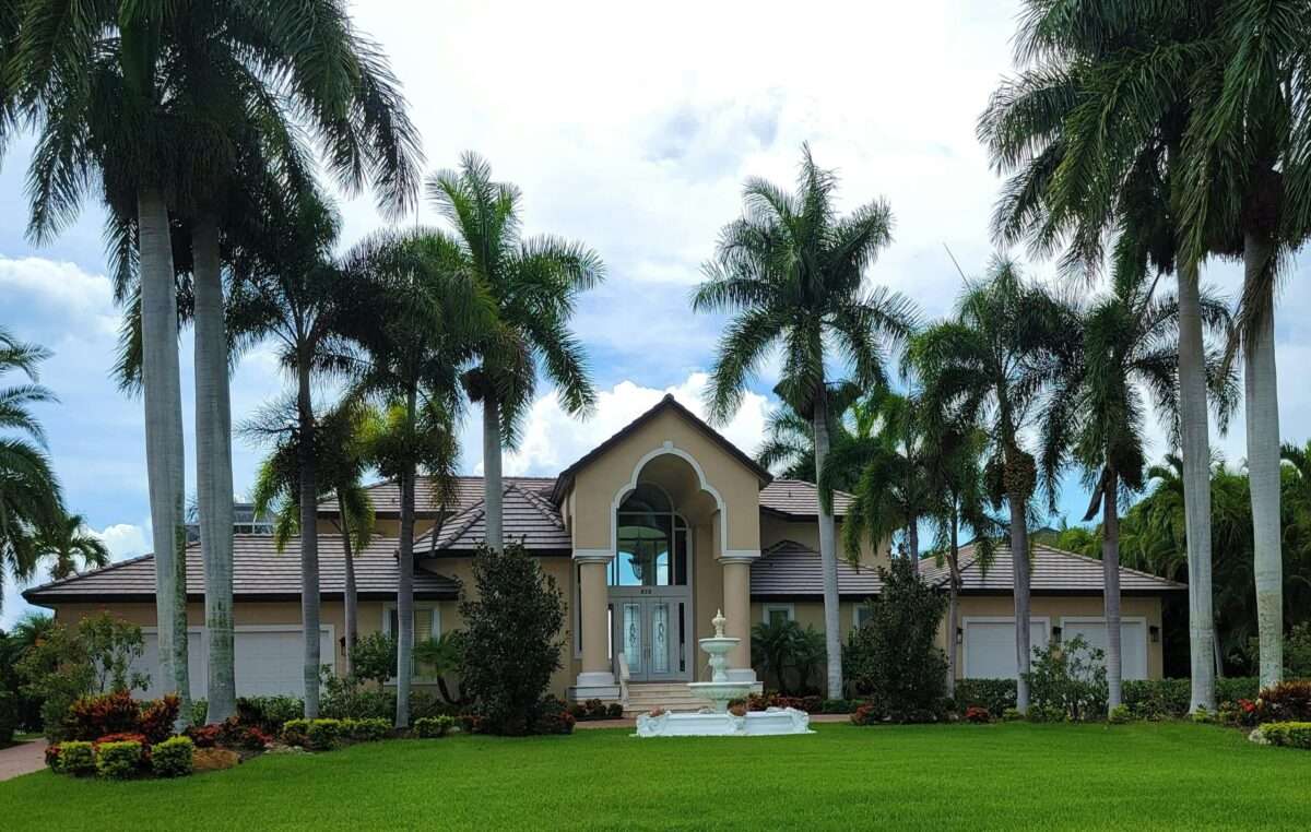 A beige tropical-style house with a centered archway, surrounded by tall palm trees and a manicured green lawn with a white fountain in front.