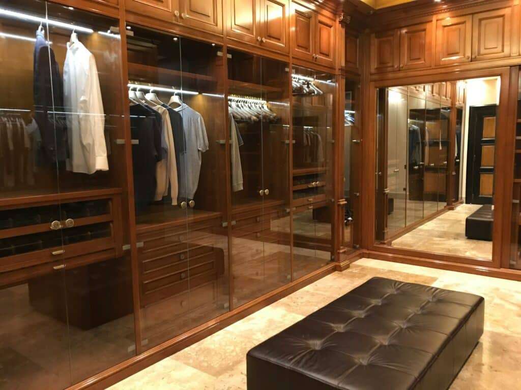 Walk-in closet with glass-front wooden wardrobes, hanging shirts and suits, under warm built-in lighting in a luxury dressing room.",