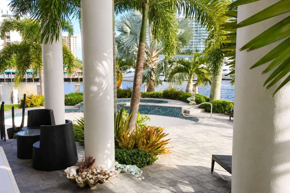 Covered waterfront patio at a Ronin Development luxury custom home featuring tropical landscaping, an elegant pool and spa, stone terrace, towering palms, and scenic Intracoastal Waterway views.