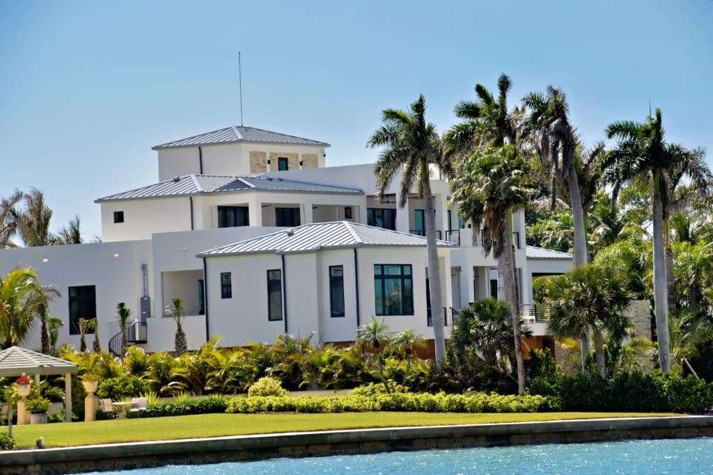 Modern luxury waterfront estate in South Florida featuring contemporary architecture, white stucco exterior, metal roofing, tropical palm trees, lush landscaping, and expansive Intracoastal views.