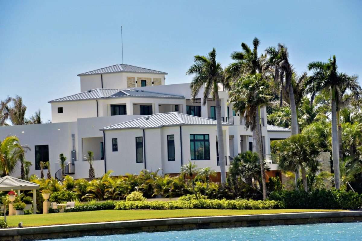 Modern luxury waterfront estate in South Florida featuring contemporary architecture, white stucco exterior, metal roofing, tropical palm trees, lush landscaping, and expansive Intracoastal views.