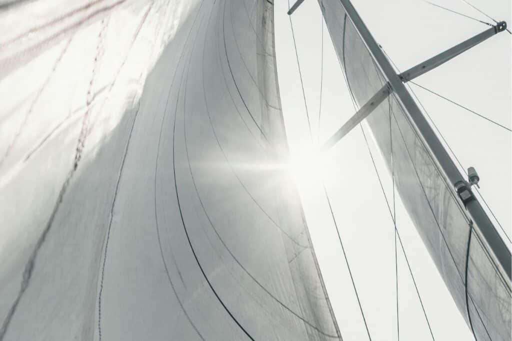 Upward view of a sailboat’s tall mast, rigging and white sails illuminated by sunlight