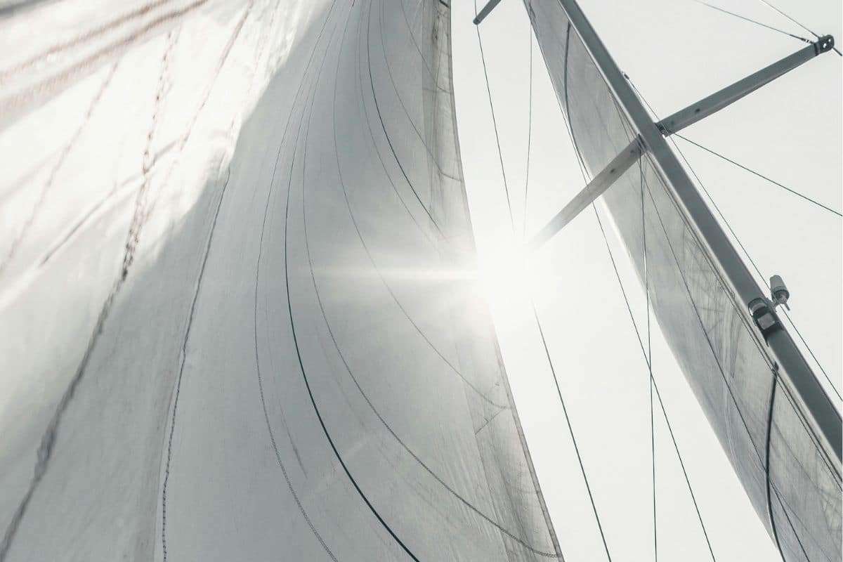 Upward view of a sailboat’s tall mast, rigging and white sails illuminated by sunlight